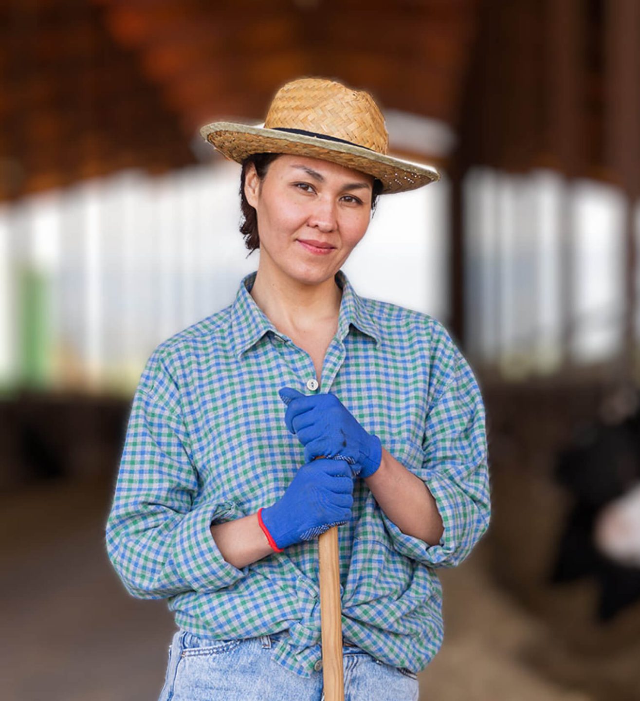 A Caucasian woman in a straw hat looking at the camera on stables background, leaning on a shovel